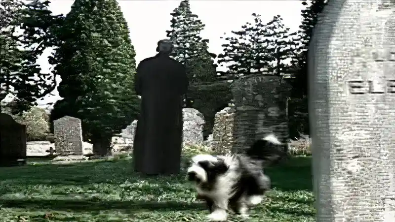Clergyman standing in a graveyard as a dog runs past gravestones in the Free As A Bird video
