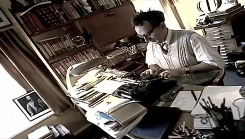 Man typing at a desk surrounded by papers and books